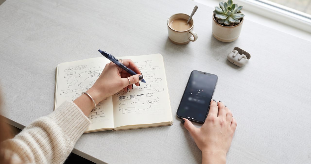 ノートに手書きでアイデアをまとめながら、隣に置いたスマートフォンを確認している人の手元