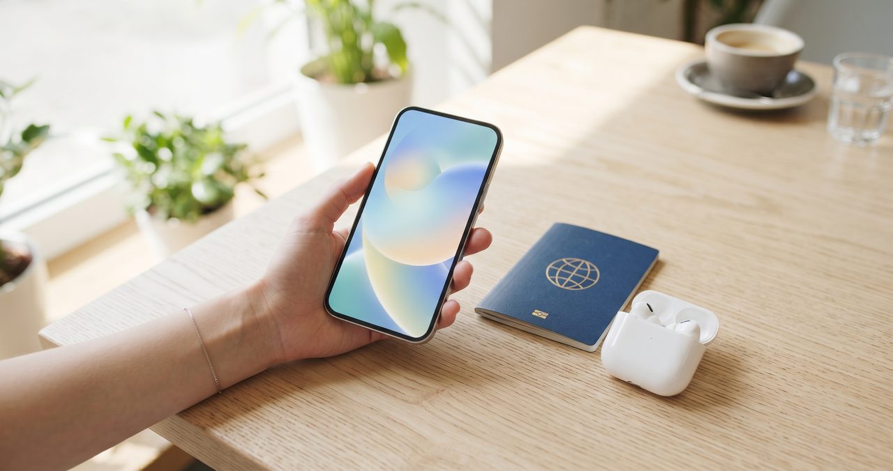 窓際のカフェでスマートフォンを手に持ち画面を確認する人物の手元、隣にパスポートが置かれ海外でもそのまま使える様子を表した写真