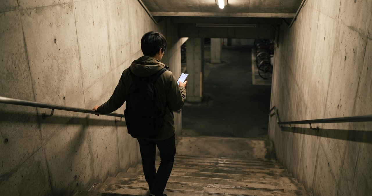 薄暗い地下駐輪場へ続くコンクリートの階段を下りる人物の後ろ姿を引きで撮影した、電波環境が変わる場面をイメージした写真