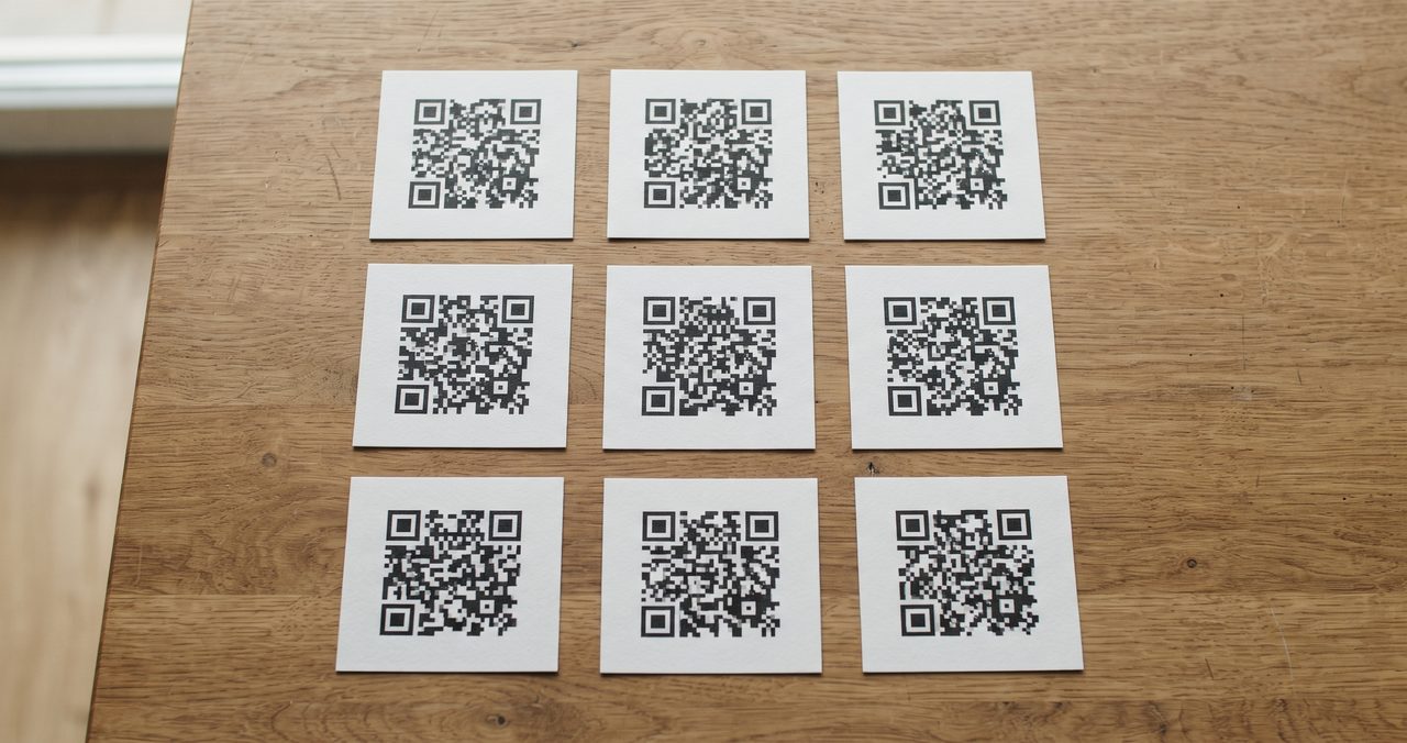 白黒のモザイク状パターンが印刷された正方形のカードが木製テーブルに並べられている俯瞰写真、QRコードを複数まとめて作成するイメージ